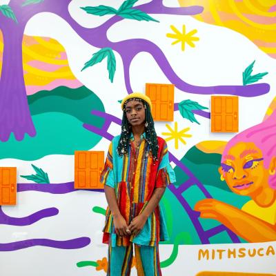 Photo of local artist Mithsuca Berry wearing top and pants with a multi-colored striped pattern and standing in front of a mural they created that has many colors and depicts people making their way across bridges, around trees, and more, and also features three-dimensional orange doors